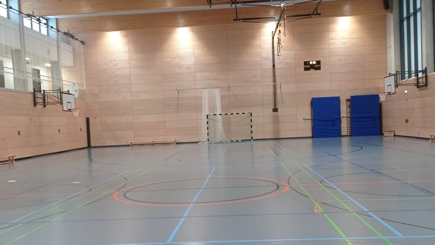Leere Sporthalle mit Spielfeldmarkierungen, Handballtor, Basketballkörben und Turnmatten an der Wand